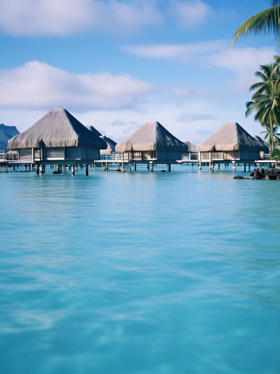 Overwater bungalows with thatched roofs on stilts over turquoise water under a blue sky, palm trees to the right.