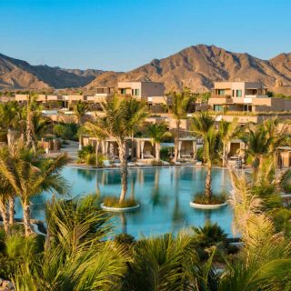 Desert resort with palm trees, circular pools, and modern villas set against rocky mountains under a blue sky.