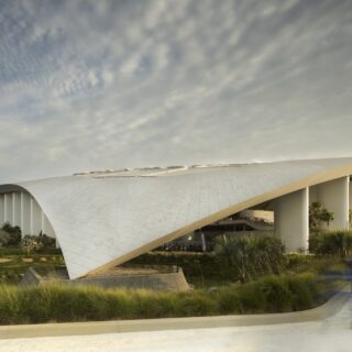 Modern curved white pavilion with open sides, surrounded by greenery and people walking along a path, under a cloudy sky.