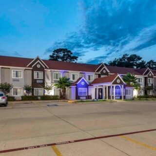 Twilight exterior of a long, two-story residential building with a central blue-lit entrance canopy and palm trees, cars parked in the lot several meters away.