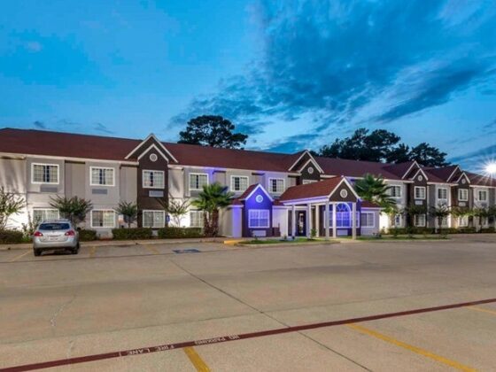 Twilight exterior of a long, two-story residential building with a central blue-lit entrance canopy and palm trees, cars parked in the lot several meters away.