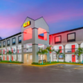 Two-story hotel with a white exterior, red doors, palm trees, and a pink sunset sky in the background.
