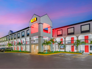Two-story hotel with a white exterior, red doors, palm trees, and a pink sunset sky in the background.