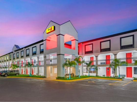Two-story hotel with a white exterior, red doors, palm trees, and a pink sunset sky in the background.