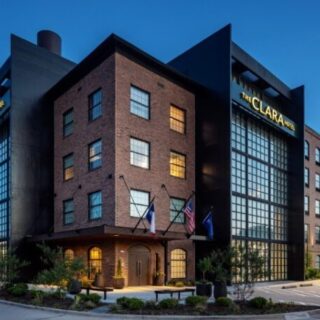 Exterior view of The Clara Hotel, a brick building with large glass atriums and flags at the entry at dusk