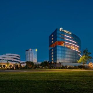 Night exterior of a modern office complex, featuring a curved glass tower labeled The Crescent and a glowing lawn sculpture.