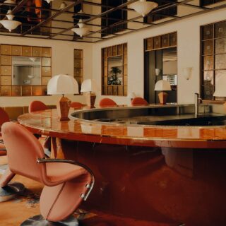 Warm-toned art deco hotel bar with a curved orange marble counter and coral chairs, mirrored panels, and globe lamps.