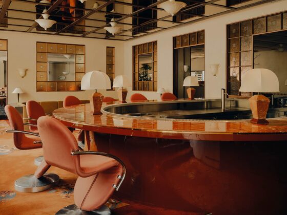 Warm-toned art deco hotel bar with a curved orange marble counter and coral chairs, mirrored panels, and globe lamps.