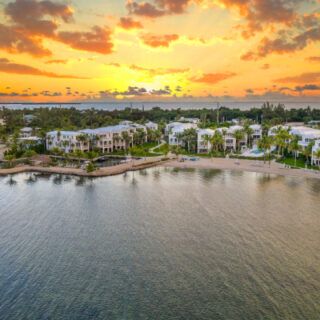 Sunset over a coastal neighborhood with white waterfront villas, palm trees, and calm water along a rocky shoreline.