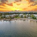 Aerial view of a beachfront resort at sunset, white buildings lining a grassy shoreline with palm trees and calm water.