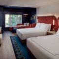 Hotel room with two large white beds, red upholstered headboards, and a seating area by a window; blue accent wall and patterned rug.