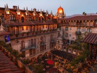 Sunset view of a Spanish-style hotel courtyard with ivy-covered balconies, arched windows, terracotta roofs, and string lights across a central plaza.