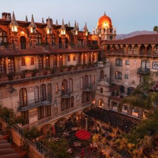 Sunset view of a Spanish-style hotel courtyard with ivy-covered balconies, arched windows, terracotta roofs, and string lights across a central plaza.