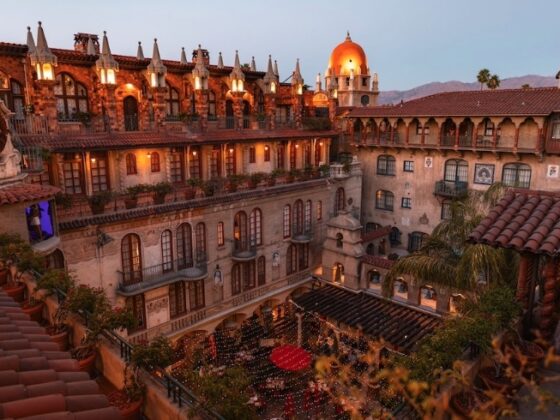 Sunset view of a Spanish-style hotel courtyard with ivy-covered balconies, arched windows, terracotta roofs, and string lights across a central plaza.