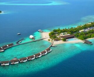 Tropical island resort with a ring of overwater bungalows around a central lagoon, connected by wooden walkways; turquoise water and palm trees.