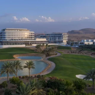 Modern resort buildings overlook a landscaped golf course with lagoons and palm trees, desert hills in the background.