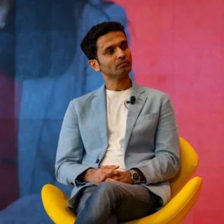 Man in a light blue blazer seated on a bright yellow chair, hands clasped, with a lapel microphone for a talk.