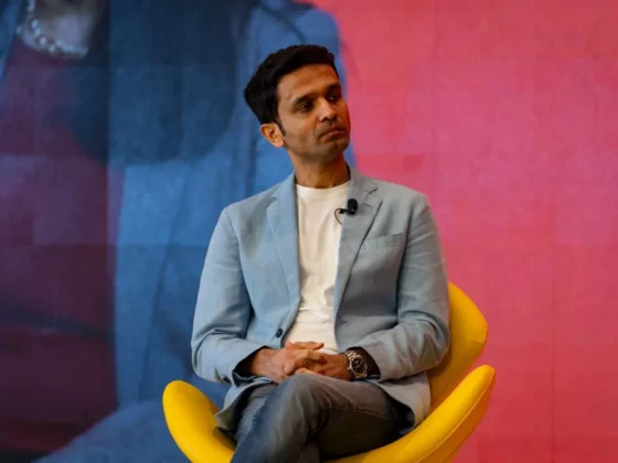 Man in a light blue blazer seated on a bright yellow chair, hands clasped, with a lapel microphone for a talk.