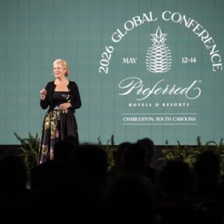 Speaker on a stage addressing an audience at the 2026 Global Conference (May 12–14) for Preferred Hotels & Resorts, in Charleston, South Carolina; backdrop includes the conference logo and branding, with silhouettes of attendees in the foreground.