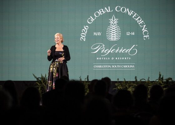 Speaker on a stage addressing an audience at the 2026 Global Conference (May 12–14) for Preferred Hotels & Resorts, in Charleston, South Carolina; backdrop includes the conference logo and branding, with silhouettes of attendees in the foreground.
