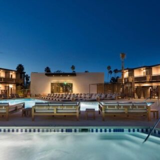 Evening view of a hotel pool deck with lounge chairs, two pools, and palm trees against a blue sky.