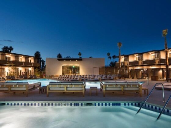 Evening view of a hotel pool deck with lounge chairs, two pools, and palm trees against a blue sky.