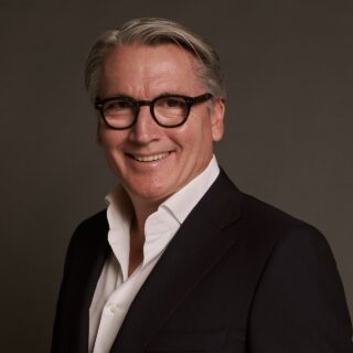 Smiling man in a dark blazer and white shirt, wearing round black glasses, posed against a brown studio backdrop.