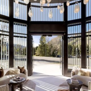 Open glass doors reveal a sunlit lobby with curved windows, hanging pendant lights, and circular seating facing a mountain view outside climber trees and road ahead.