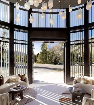 Open glass doors reveal a sunlit lobby with curved windows, hanging pendant lights, and circular seating facing a mountain view outside climber trees and road ahead.