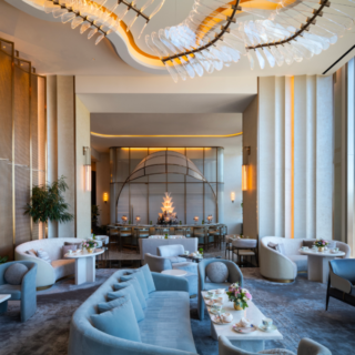 Luxurious hotel lounge with pale blue sofas, marble tables, and a dramatic ceiling chandelier overlooking city windows.