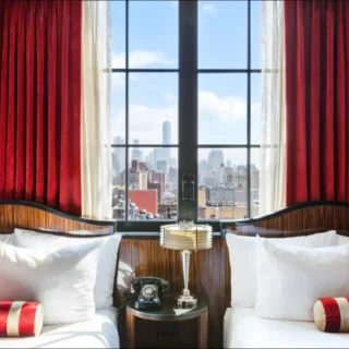 Twin beds with white linens and red bolster pillows flank a nightstand and a window view of the city skyline with red curtains framing the scene.