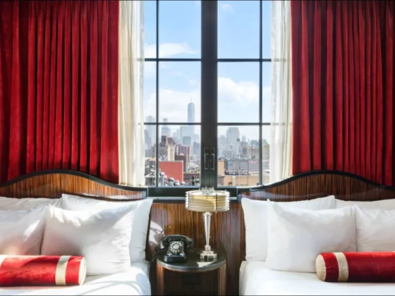 Twin beds with white linens and red bolster pillows flank a nightstand and a window view of the city skyline with red curtains framing the scene.
