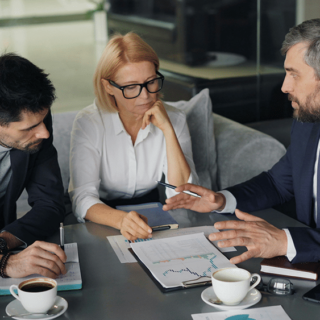 Three professionals discuss charts and notes at a conference table with coffee cups nearby.