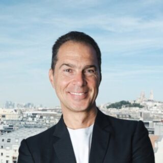 Smiling man in a dark blazer, viewed from the shoulders up, with a city skyline and blue sky in the background.