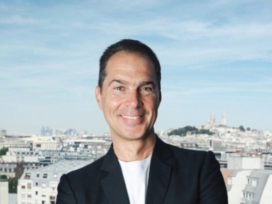 Smiling man in a dark blazer, viewed from the shoulders up, with a city skyline and blue sky in the background.