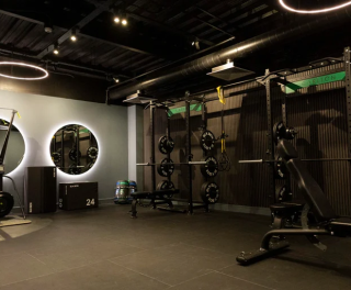 Modern gym interior with black walls, weight racks, barbells, a bench press, and circular ceiling lights.