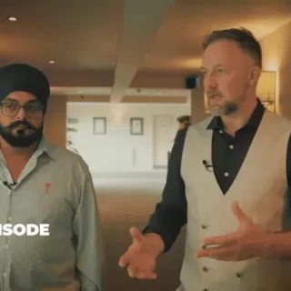 Two men in a hotel hallway: a bearded man with a turban and glasses stands beside a man in a vest who is speaking, with podcast branding text in the corner.