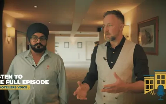 Two men in a hotel hallway: a bearded man with a turban and glasses stands beside a man in a vest who is speaking, with podcast branding text in the corner.