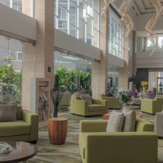 Modern hotel lobby with lime-green couches, wooden side tables, and large glass walls filled with tropical plants; bright and airy seating area. Readable distance people in background near a reception.