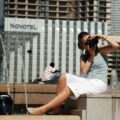 Woman with camera photographing at an urban fountain, wearing a light blue top and white shorts, with helmet on ground nearby.