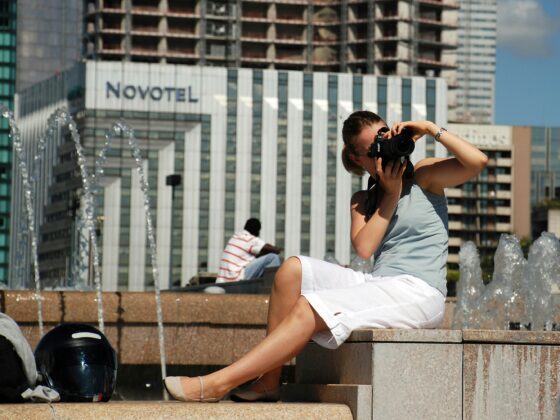 Woman with camera photographing at an urban fountain, wearing a light blue top and white shorts, with helmet on ground nearby.