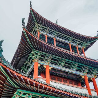 Colorful multi-tier Chinese temple roof with upturned eaves and red supporting pillars against a pale sky, architectural detail visible