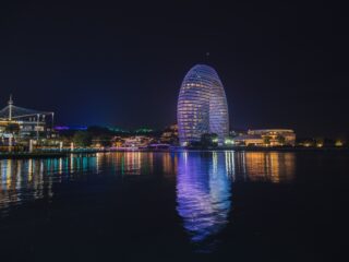 Nighttime city skyline with two glowing oval towers wrapped in blue lights by a river, reflections in the water.