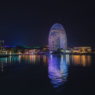 Nighttime city skyline with two glowing oval towers wrapped in blue lights by a river, reflections in the water.