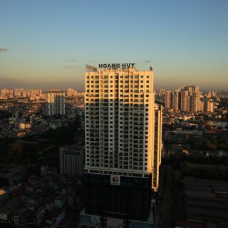 Tall white apartment building with a Hoang Huy sign on top against a sunset cityscape in the foreground.