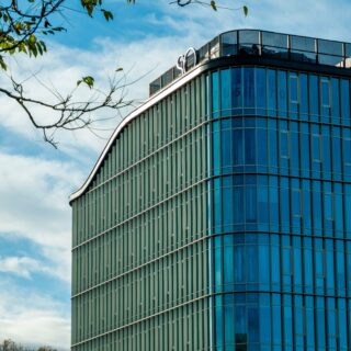 Modern glass office building with vertical green fins and blue-tinted windows under a blue sky with clouds.