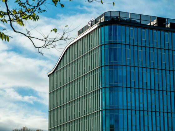 Modern glass office building with vertical green fins and blue-tinted windows under a blue sky with clouds.