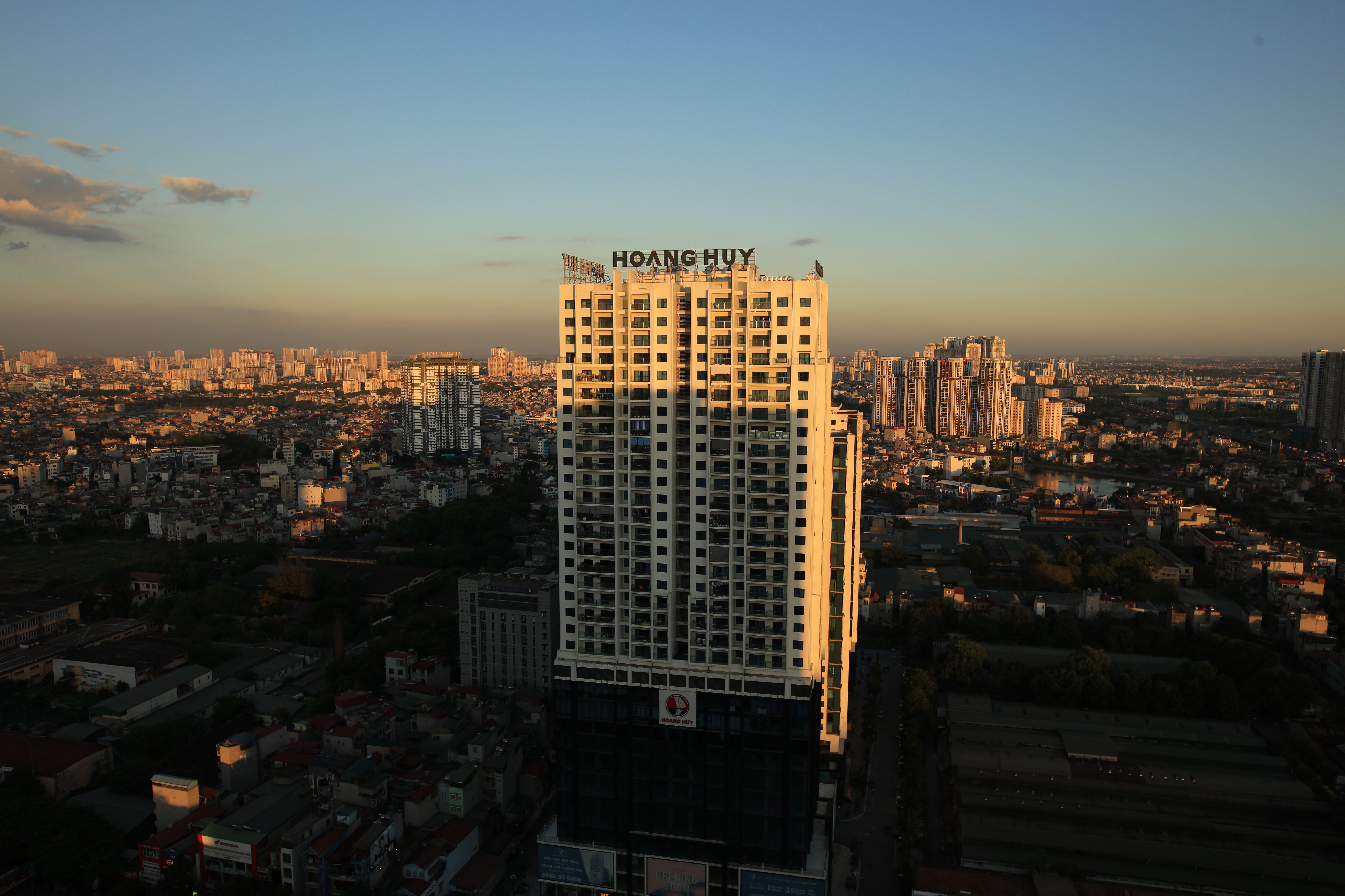 Tall white apartment building with a Hoang Huy sign on top against a sunset cityscape in the foreground.