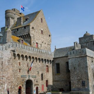 Medieval stone town hall with a crenellated tower, flags flying atop, and a blue sky. In front is a stone wall and arched doorway.