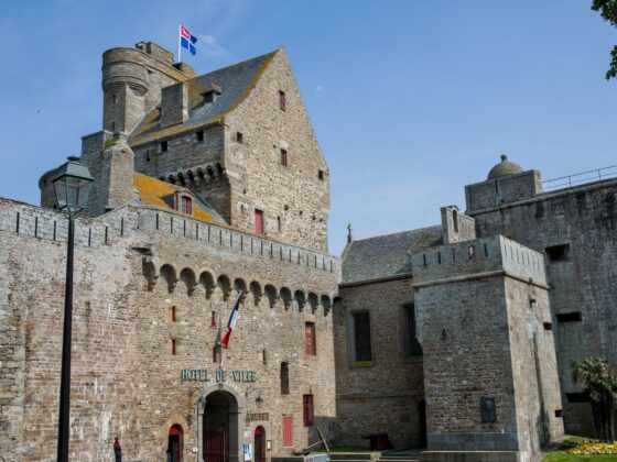 Medieval stone town hall with a crenellated tower, flags flying atop, and a blue sky. In front is a stone wall and arched doorway.